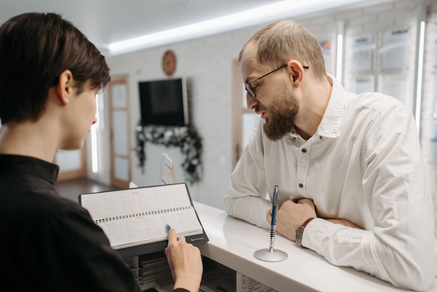 A receptionist and a patient looking at an appointment book across a clinic front desk.